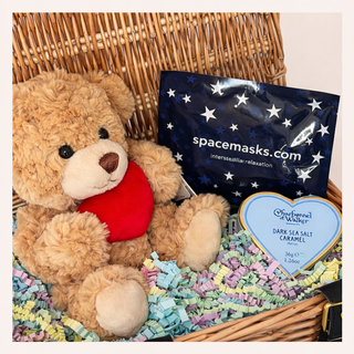 Teddy bear with a bag labeled 'spacemasks.com' and a heart-shaped chocolate bar on a woven basket.