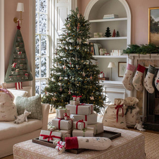 A decorated Christmas tree stands in a cozy living room with wrapped gift boxes on a table in front, stockings hanging by the fireplace, and a warm, festive interior.