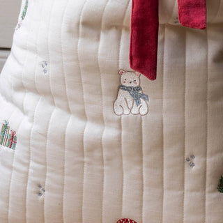 Close-up of a quilted cream fabric item with a small embroidered bear wearing a blue scarf, a red velvet tie at the top, and subtle holiday motifs scattered across the surface.