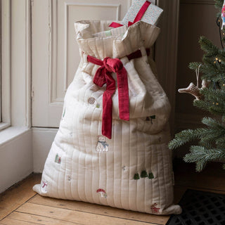 A large quilted Christmas sack in cream with embroidered woodland motifs and a red ribbon bow, standing on a wooden floor next to a Christmas tree.