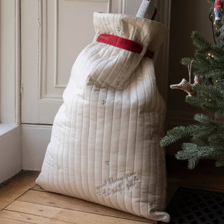 A quilted cream Christmas stocking filled with gifts, tied with a red ribbon, standing on a wooden floor next to a Christmas tree.