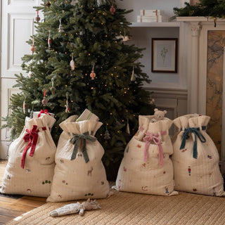 Four quilted cream holiday gift bags with festive prints and colorful ribbons, arranged on a rug in front of a decorated Christmas tree and fireplace.