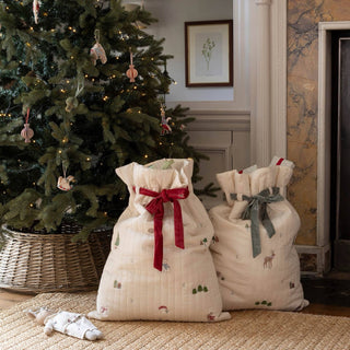 Two cream quilted Christmas stocking bags with festive prints and ribbons, placed on a woven rug in front of a decorated Christmas tree in a cozy indoor setting.