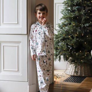 A young boy wearing a two-piece Christmas pajama set with a white base and colorful holiday motifs, standing in a doorway next to a decorated Christmas tree.