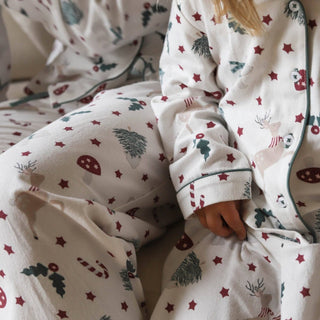 A child wearing a cozy white long-sleeve pajama set with a Christmas print, featuring red stars, green trees, candy canes, and reindeer, sitting on a bed.