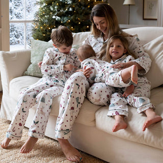 A woman and three children wearing matching white Christmas-themed pajamas with a red berry and green pine branch pattern, sitting together on a beige couch in front of a decorated Christmas tree.