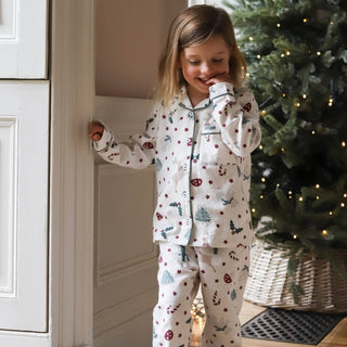 A young girl wearing a festive Christmas-themed pajama set with a white base and colorful holiday prints, standing in a doorway next to a decorated Christmas tree.