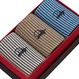 Three pairs of striped dress socks in blue, tan, and gray, presented in a red gift box. Each pair features a small embroidered dark logo on the front.