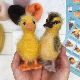 Two fluffy yellow ducklings held in a hand with a craft instruction sheet in the background.