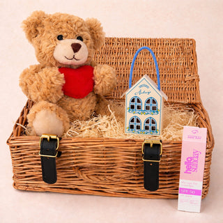 A wicker gift basket with a plush brown teddy bear holding a red heart, a small decorative house ornament, and a pink 'hello sunday' lip balm, arranged on a light beige background.