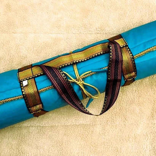A rolled turquoise blue yoga mat wrapped with decorative straps in gold, brown, and dark red, tied with a gold cord, resting on a beige textured surface.